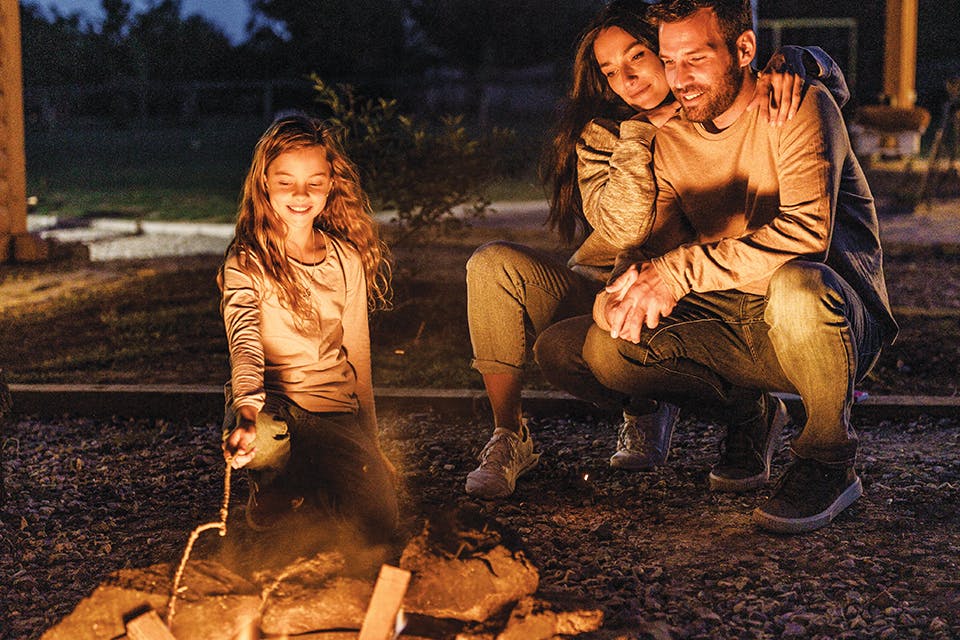 Mom, dad and daughter around a campfire outdoors (photo by iStock)
