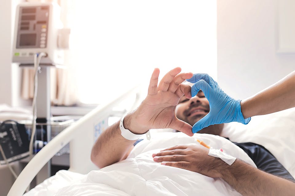 Man in hospital bed making heart with doctor’s hand (photo by iStock)